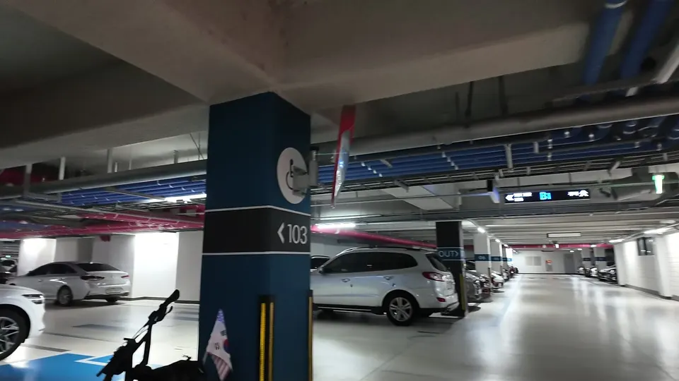 Underground car park with blue and pink ceiling pipes, a dark blue pillar and my kick scooter leaning by the disabled bay