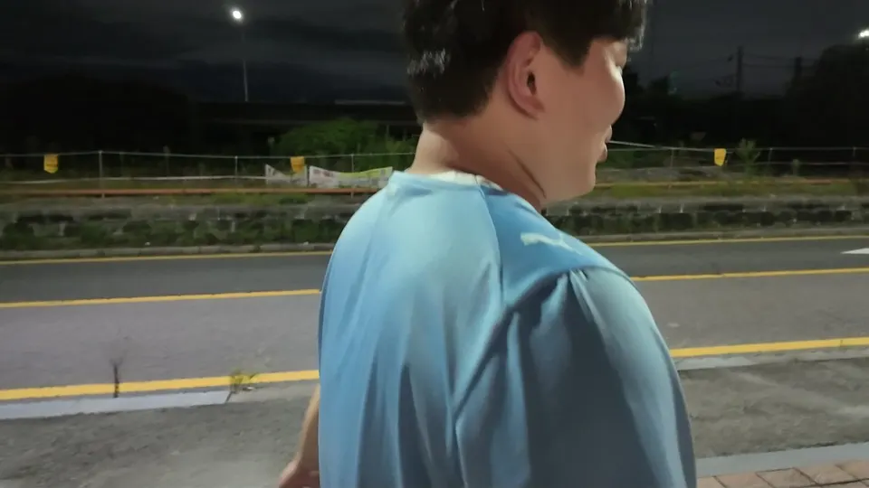 A friend in a light blue football shirt seen from behind, walking along the shoulder of a road with a double yellow line