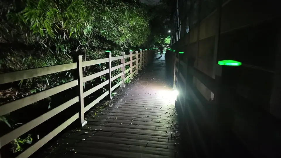 Damp wooden walkway with leaves on the boards, green post lights on both sides, a figure far ahead under a streetlight