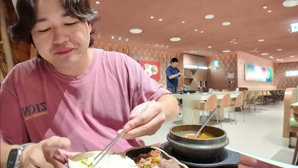 Eddie eating rice and bulgogi with chopsticks next to a boiling stone pot of stew in an empty Incheon food court