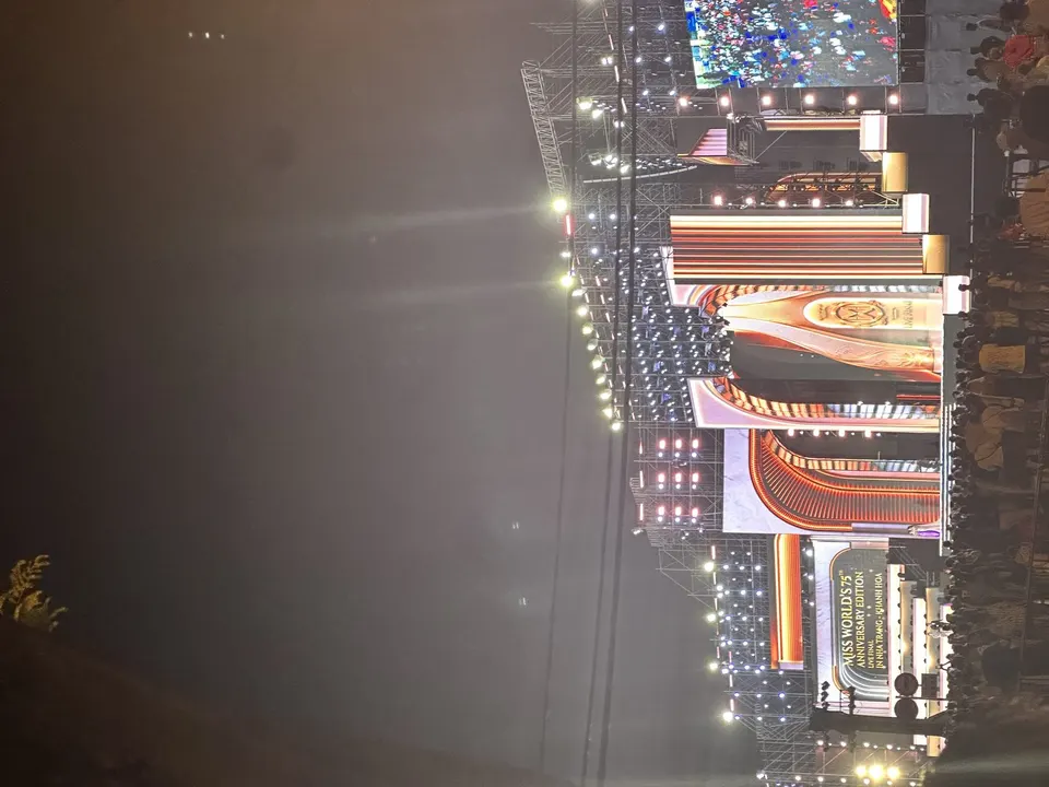Distant outdoor stage glowing through evening haze with Miss World 75th Anniversary signage and crowds in front