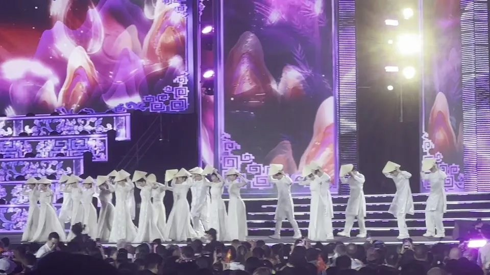 Rows of performers in white ao dai and conical hats dancing on the stage steps above a crowd holding phones