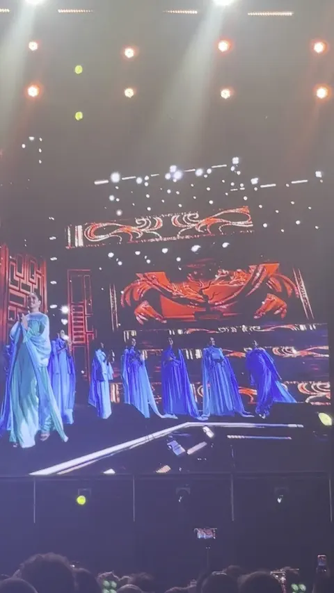 A singer in green performing in front of dancers in long blue capes under red stage lighting