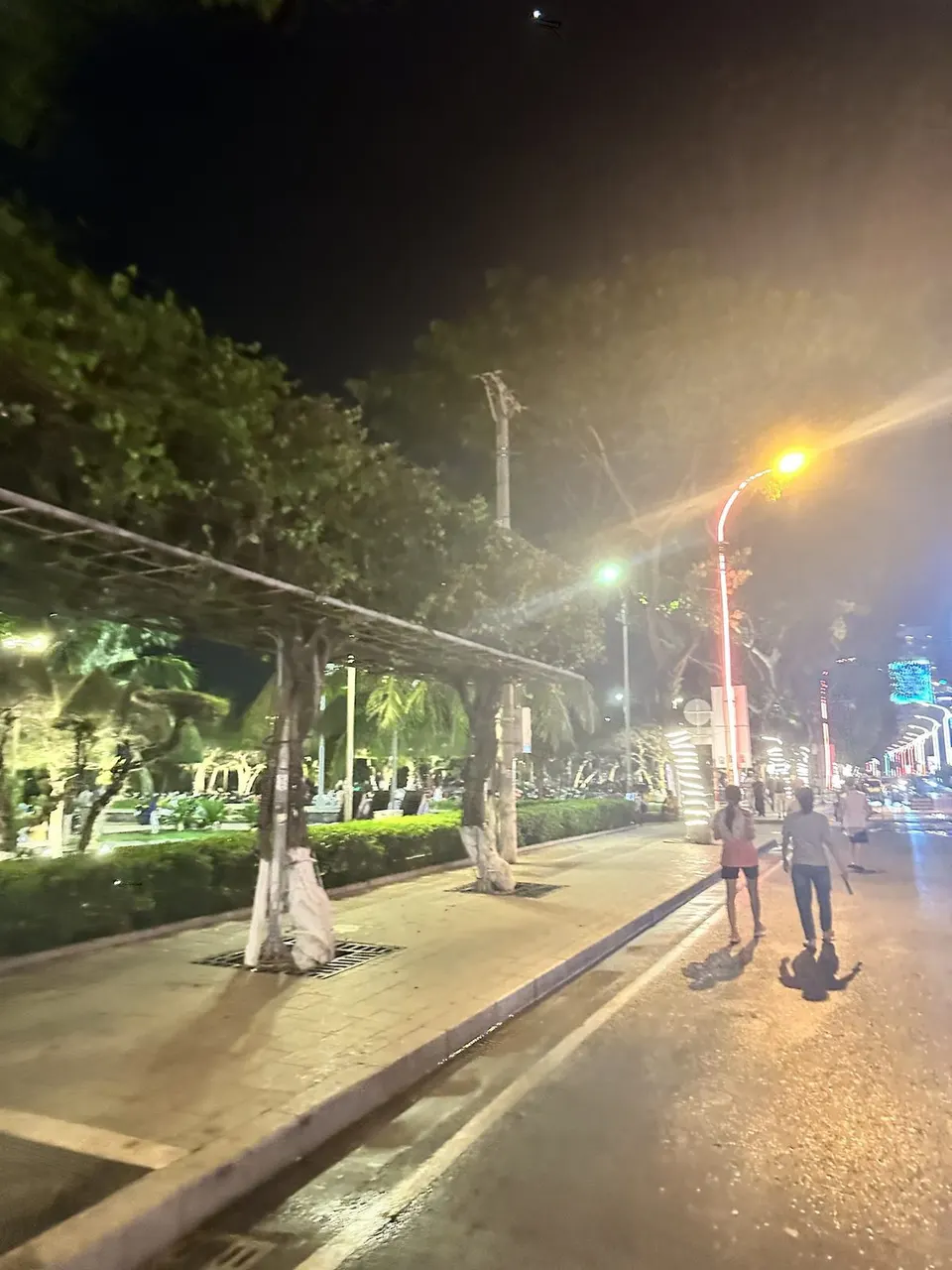 Two people walking away down a night sidewalk beside a lit park, event lights in the distance