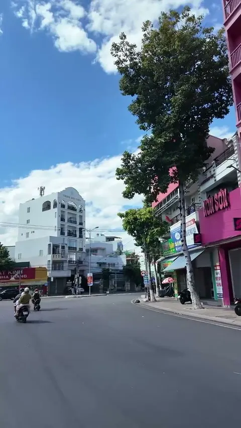 City street with a bright pink shopfront on the right, shop signs, trees and a few riders in the distance