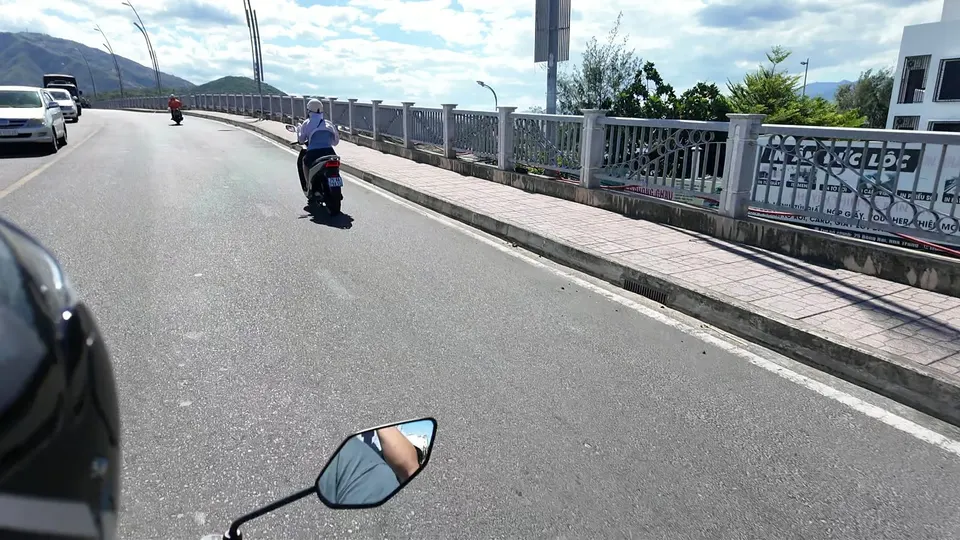 View over the scooter mirror on a bridge with new railings, a rider in a white sun jacket ahead