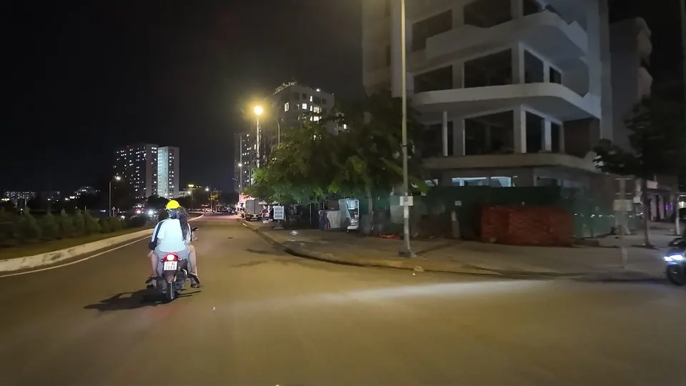 Two people on a scooter ahead on a dark road, passenger in a yellow helmet, unfinished building on the right