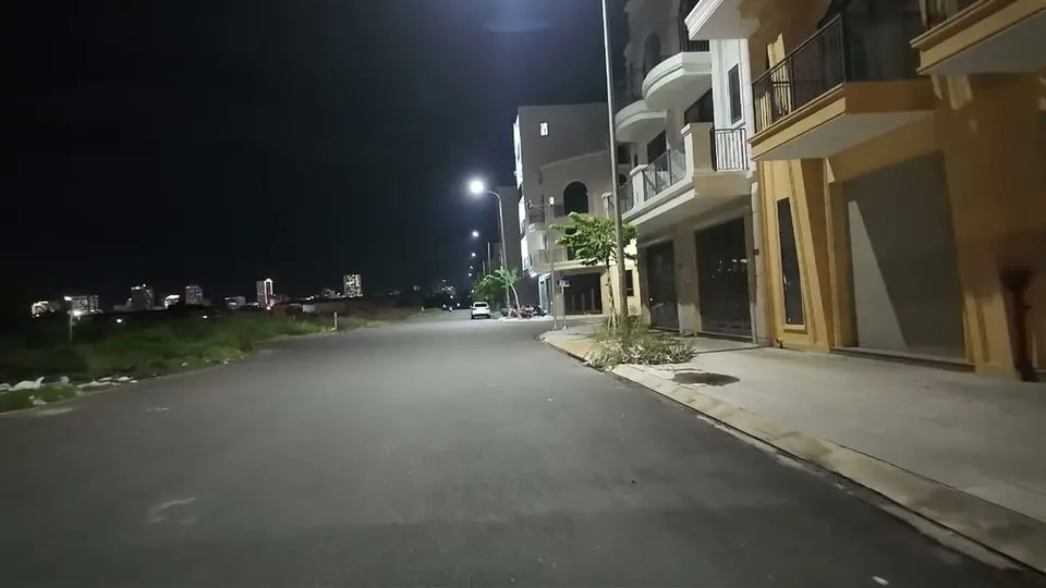 Empty new street at night lined by tall unlit shophouses, dark open lots on the other side